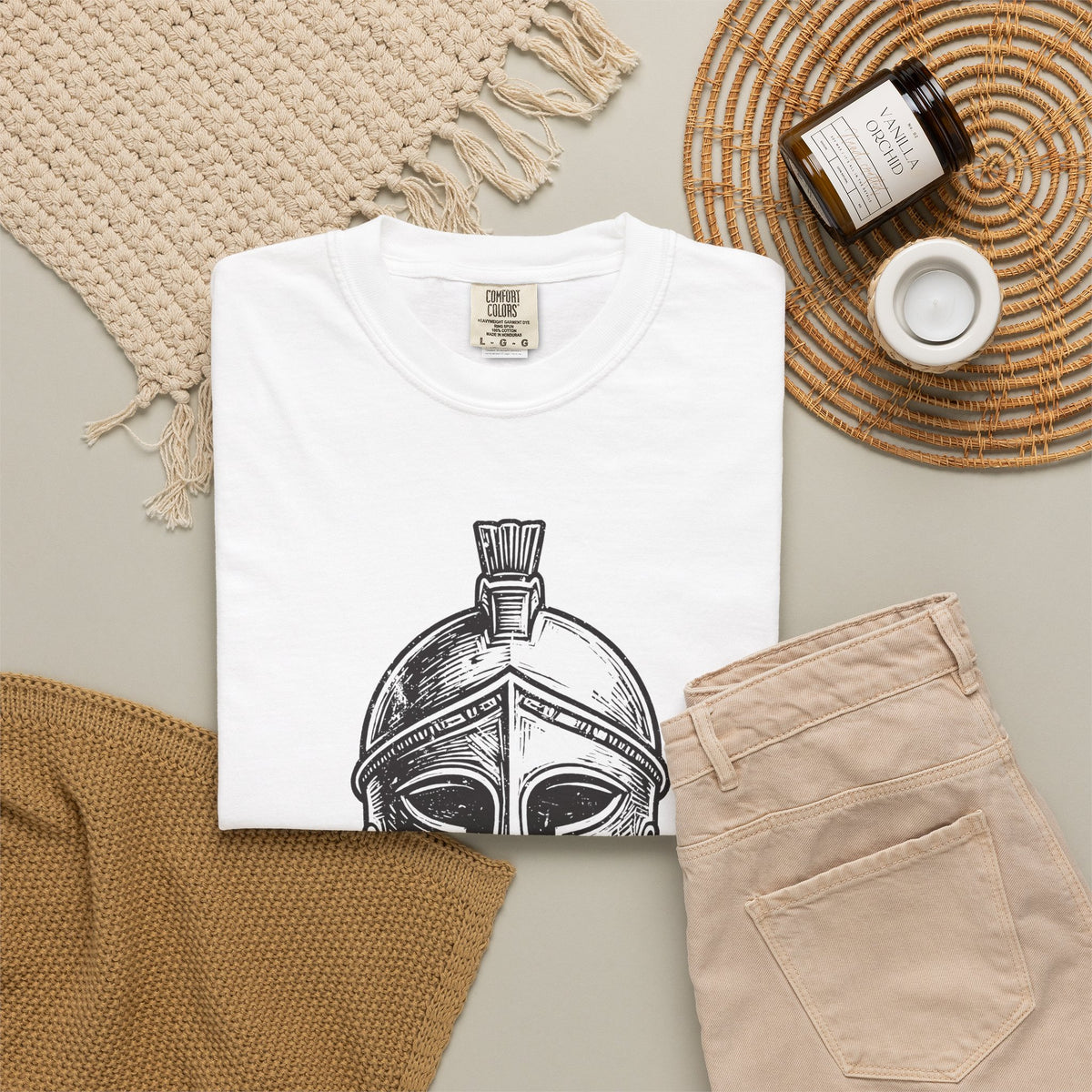 Folded white T-shirt with a bold black woodcut Corinthian helmet graphic, styled in a flat lay with beige textiles, trousers, and a candle.